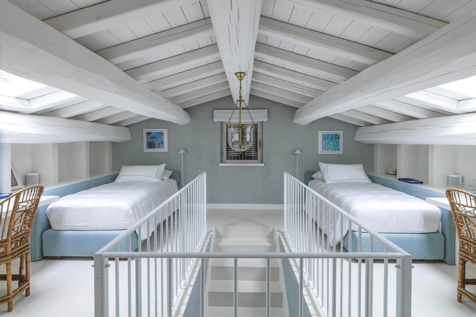 Bright and airy loft bedroom featuring two twin beds with light blue headboards, white bedding, and a high vaulted ceiling with exposed beams. Natural light floods the space through skylights, and decorative accents include framed artwork and a modern light fixture. Ideal for a serene and stylish retreat. | Only on SopranoVillas