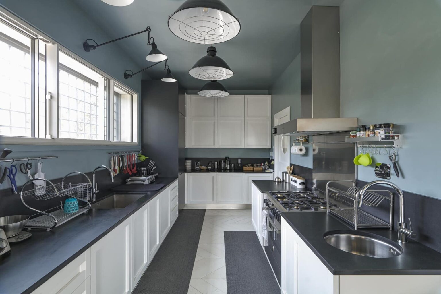 Modern kitchen interior featuring sleek white cabinetry, dark countertops, and industrial-style lighting. The space is brightened by large windows and showcases a functional layout with stainless steel appliances. | Only on SopranoVillas