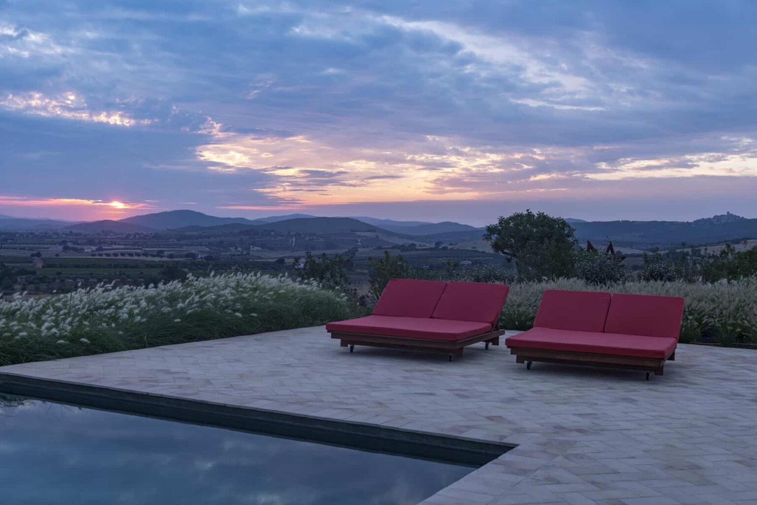 Modern outdoor lounge area with two red sun loungers beside a swimming pool, overlooking a scenic sunset and rolling hills. | Only on SopranoVillas