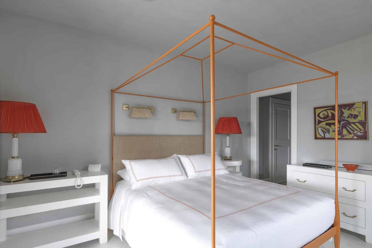 Modern bedroom featuring a stylish four-poster bed with an orange frame, complemented by white bedding. The room includes elegant bedside lamps with red shades and a minimalist white nightstand, creating a bright and inviting atmosphere. | Only on SopranoVillas