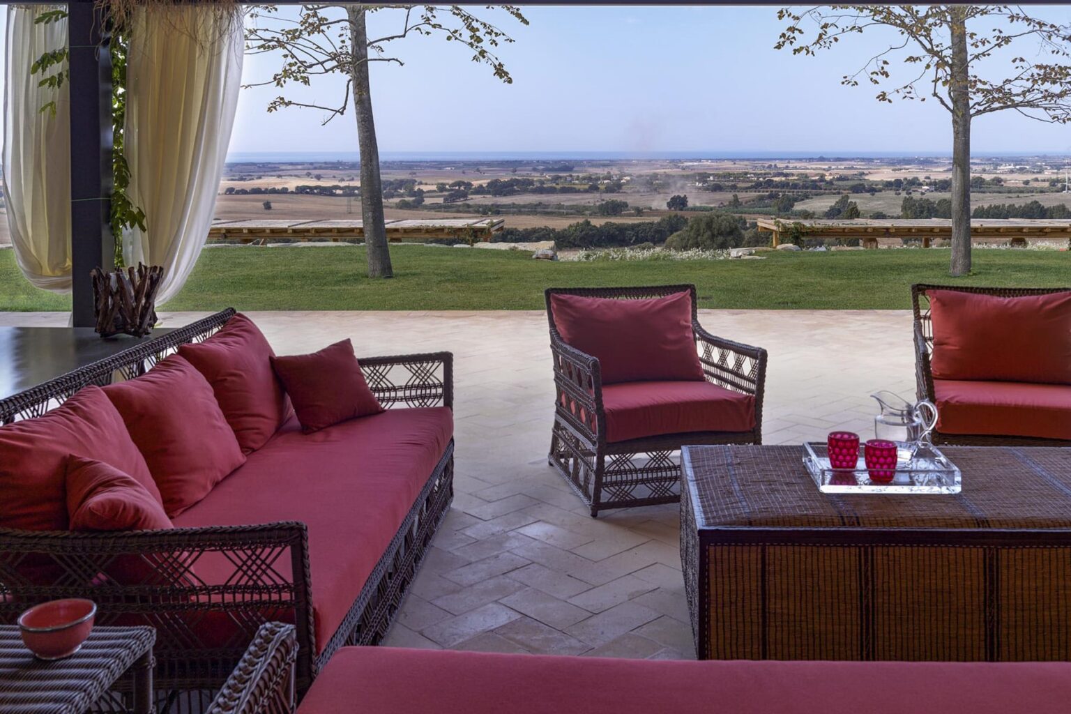 Stylish outdoor lounge area featuring comfortable red cushions on wicker furniture, overlooking a scenic landscape with trees and distant hills. Ideal for relaxation and enjoying nature. | Only on SopranoVillas