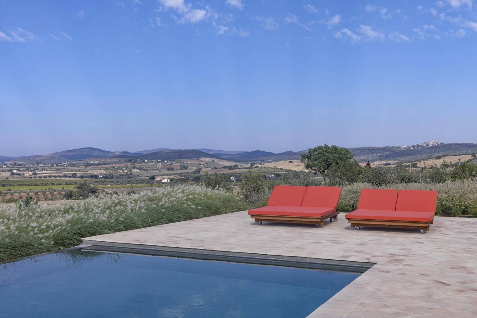Relaxing outdoor space featuring two red lounge chairs by a swimming pool, with scenic views of rolling hills and a clear blue sky. Ideal for vacation or leisure activities. | Only on SopranoVillas