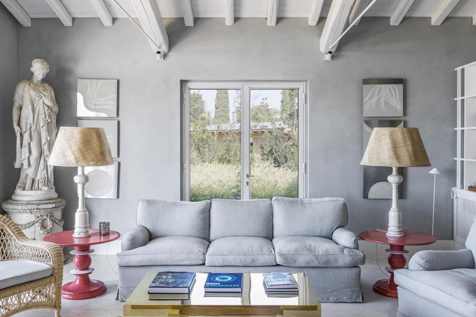 Modern living room featuring a light gray sofa, red side tables, and stylish lamps, with large windows overlooking greenery and a decorative statue in the corner. | Only on SopranoVillas