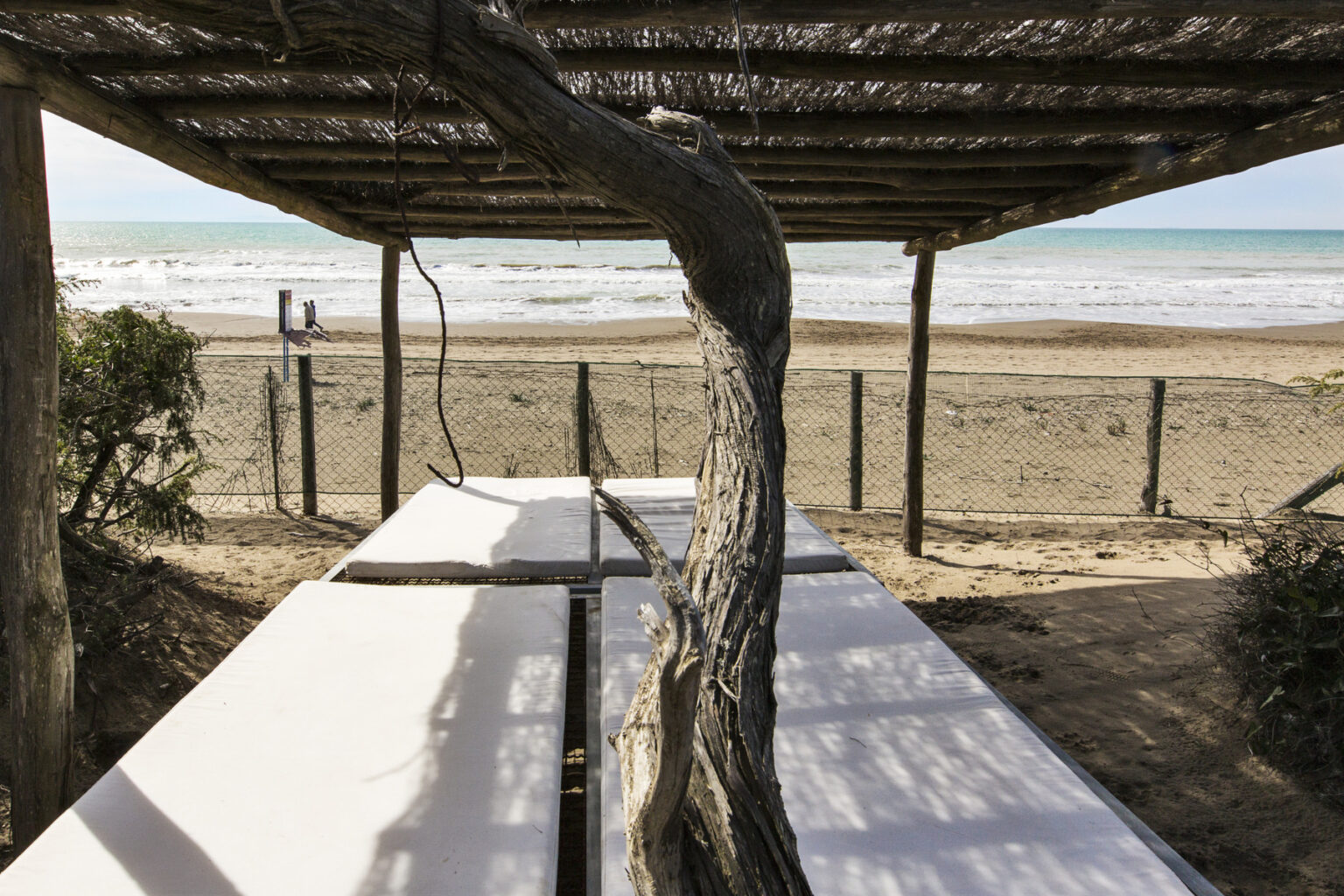 Beachside relaxation area featuring white lounge beds under a wooden pergola, with a view of the ocean and sandy shore in the background. | Only on SopranoVillas