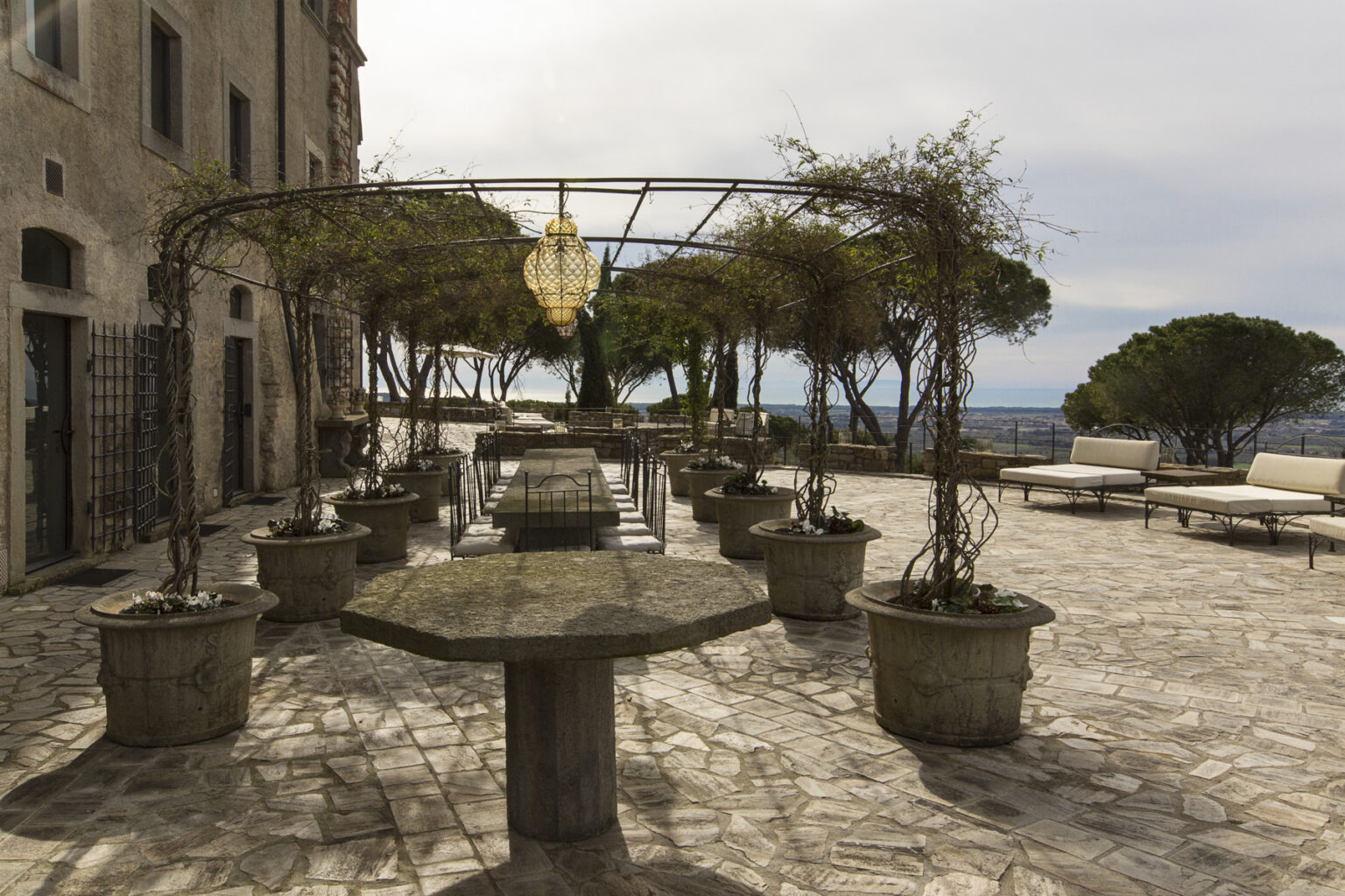 Outdoor terrace featuring stone tables and potted plants, with a decorative chandelier and trees in the background, offering a scenic view. Ideal for gatherings or events in a serene setting. | Only on SopranoVillas