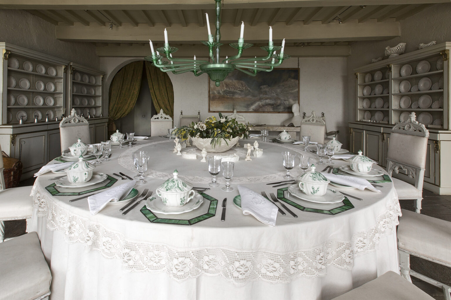 Elegant dining room featuring a round table set with fine china, green accents, and a floral centerpiece, surrounded by white chairs and a chandelier. Shelves in the background display additional dishware, enhancing the sophisticated atmosphere. | Only on SopranoVillas