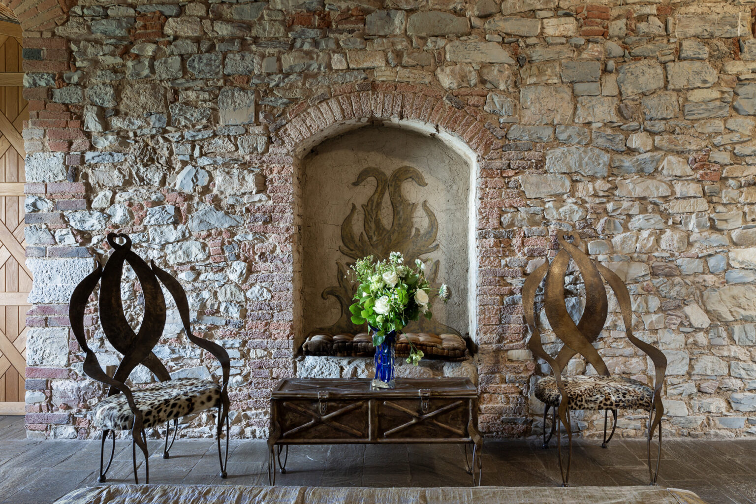 Rustic interior design featuring a stone wall, two artistic metal chairs with leopard print cushions, and a wooden coffee table adorned with a blue vase of white flowers, creating a charming and cozy atmosphere. | Only on SopranoVillas