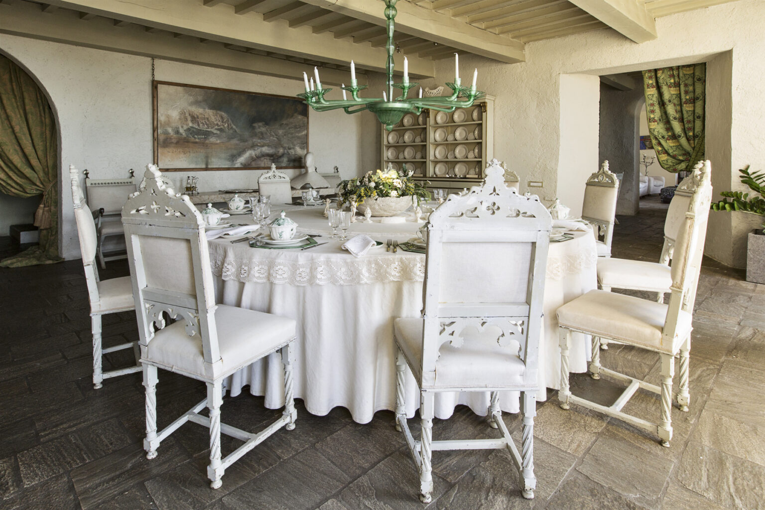 Elegant dining room featuring a round table set with white tableware, surrounded by ornate white chairs. A green chandelier hangs above, complementing the sophisticated decor and natural light coming from large windows. | Only on SopranoVillas
