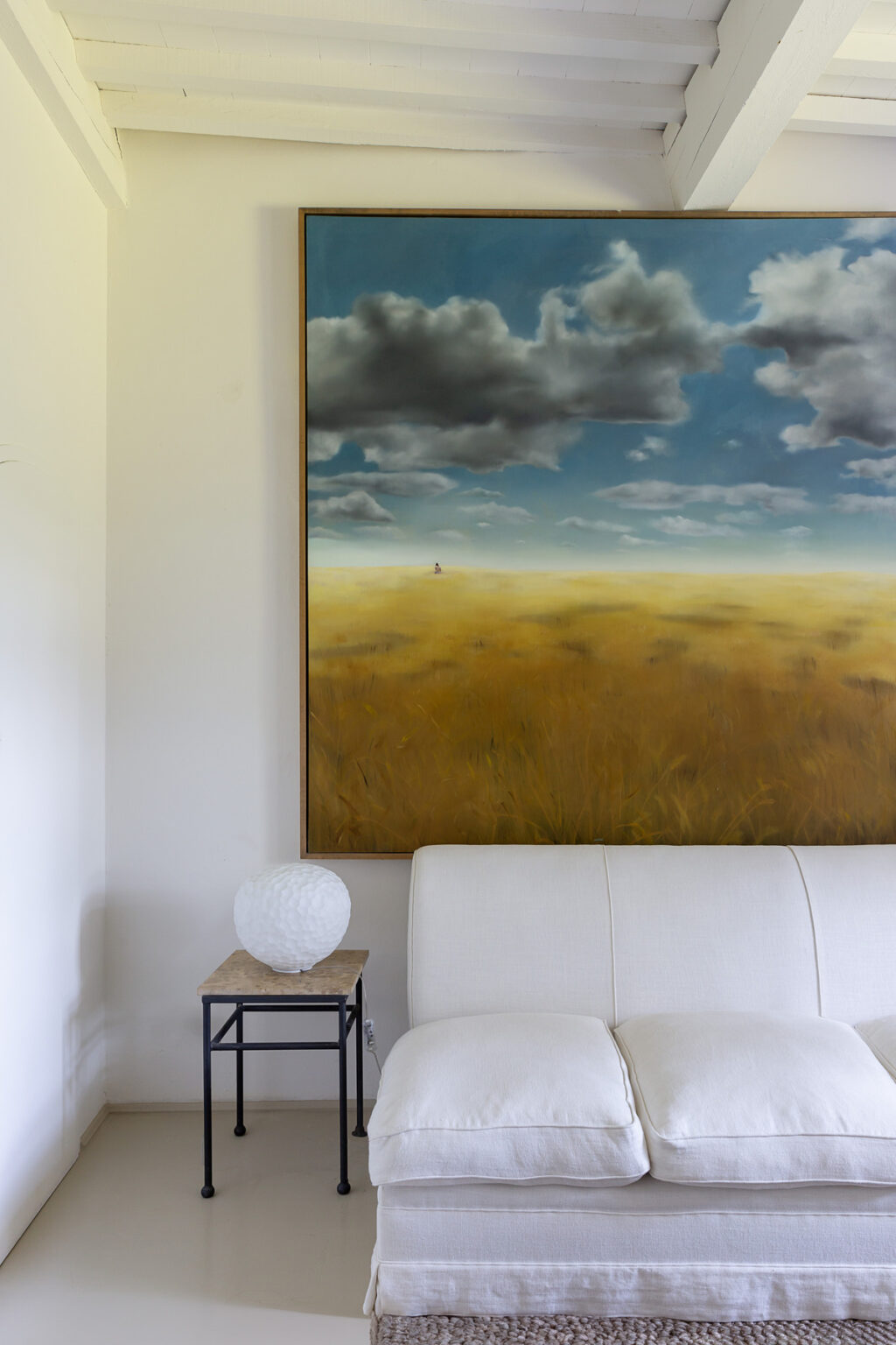 A serene living room featuring a large landscape painting of a golden field under a cloudy blue sky, complemented by a modern white sofa and a decorative lamp. | Only on SopranoVillas