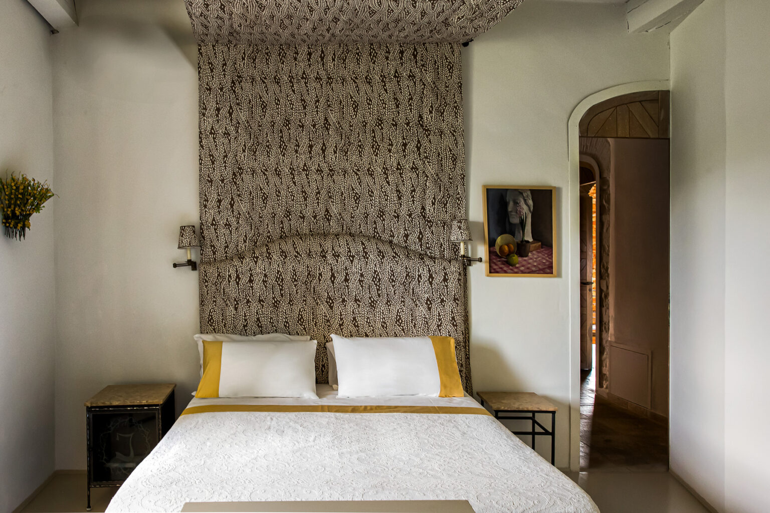 Cozy bedroom featuring a stylish bed with white and yellow bedding, a patterned accent wall, and decorative wall art. The room is softly lit, creating a warm atmosphere, with a glimpse of an archway leading to another space. | Only on SopranoVillas