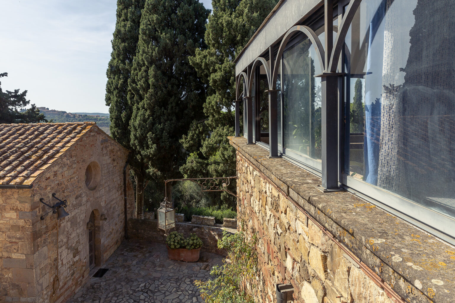 Scenic view of a rustic stone building beside a modern structure with large windows, surrounded by lush greenery and cypress trees in a tranquil outdoor setting. | Only on SopranoVillas