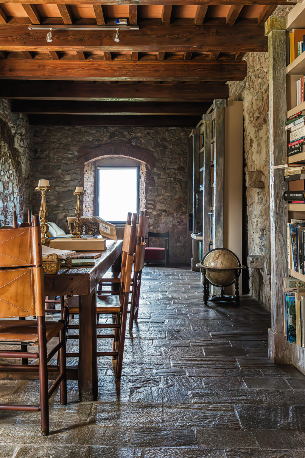 Cozy reading room featuring a wooden dining table, leather chairs, a vintage globe, and stone walls, illuminated by natural light from a window. Bookshelves line the walls, creating an inviting atmosphere for study and relaxation. | Only on SopranoVillas