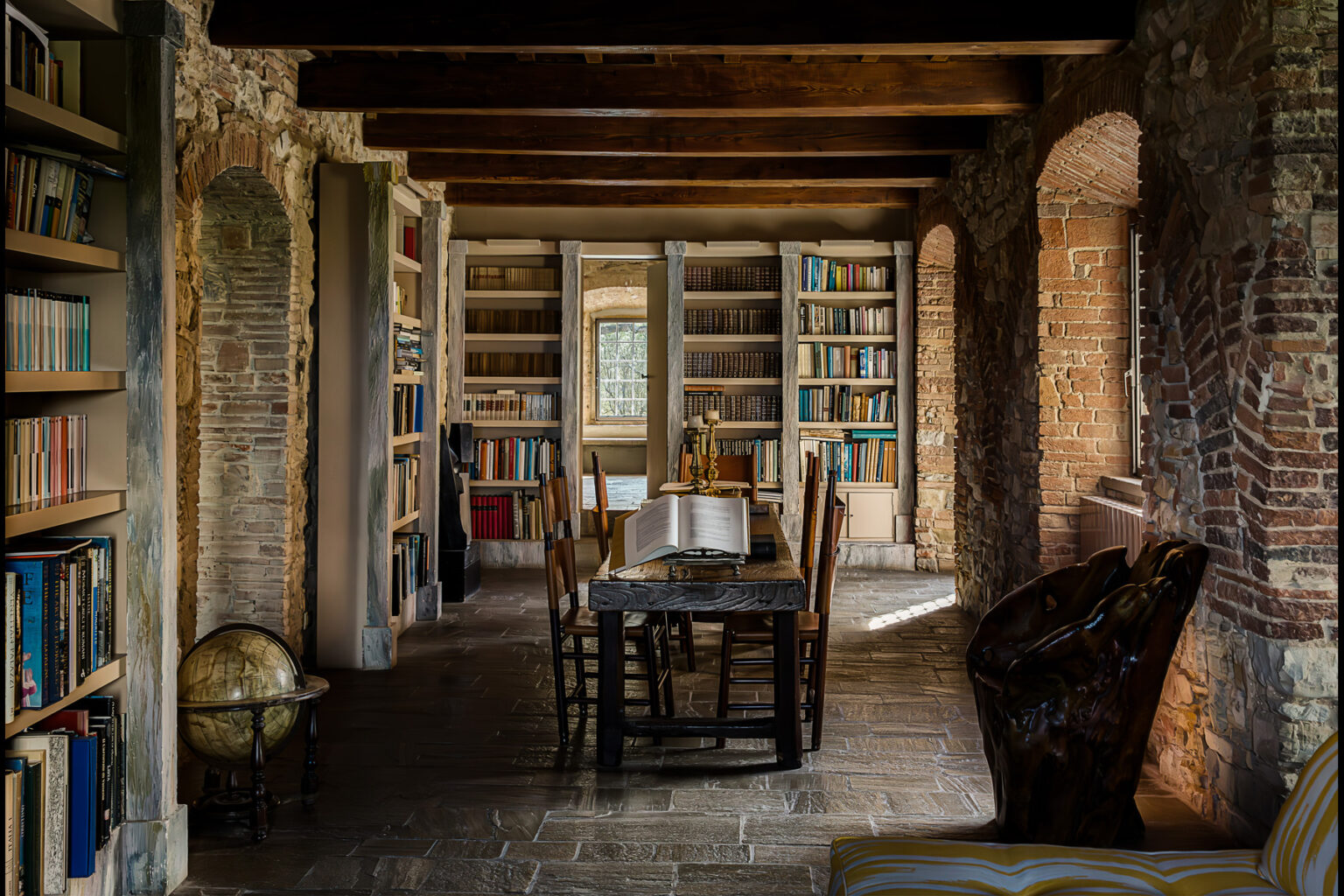 Cozy library interior featuring wooden bookshelves filled with books, a rustic table surrounded by chairs, and a vintage globe, all set against exposed brick walls and wooden beams. | Only on SopranoVillas