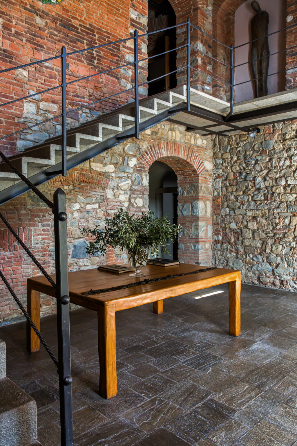 Interior view of a modern home featuring a wooden table with a vase of greenery, set against a backdrop of textured stone walls and a staircase. The design combines rustic and contemporary elements, creating a warm and inviting atmosphere. | Only on SopranoVillas