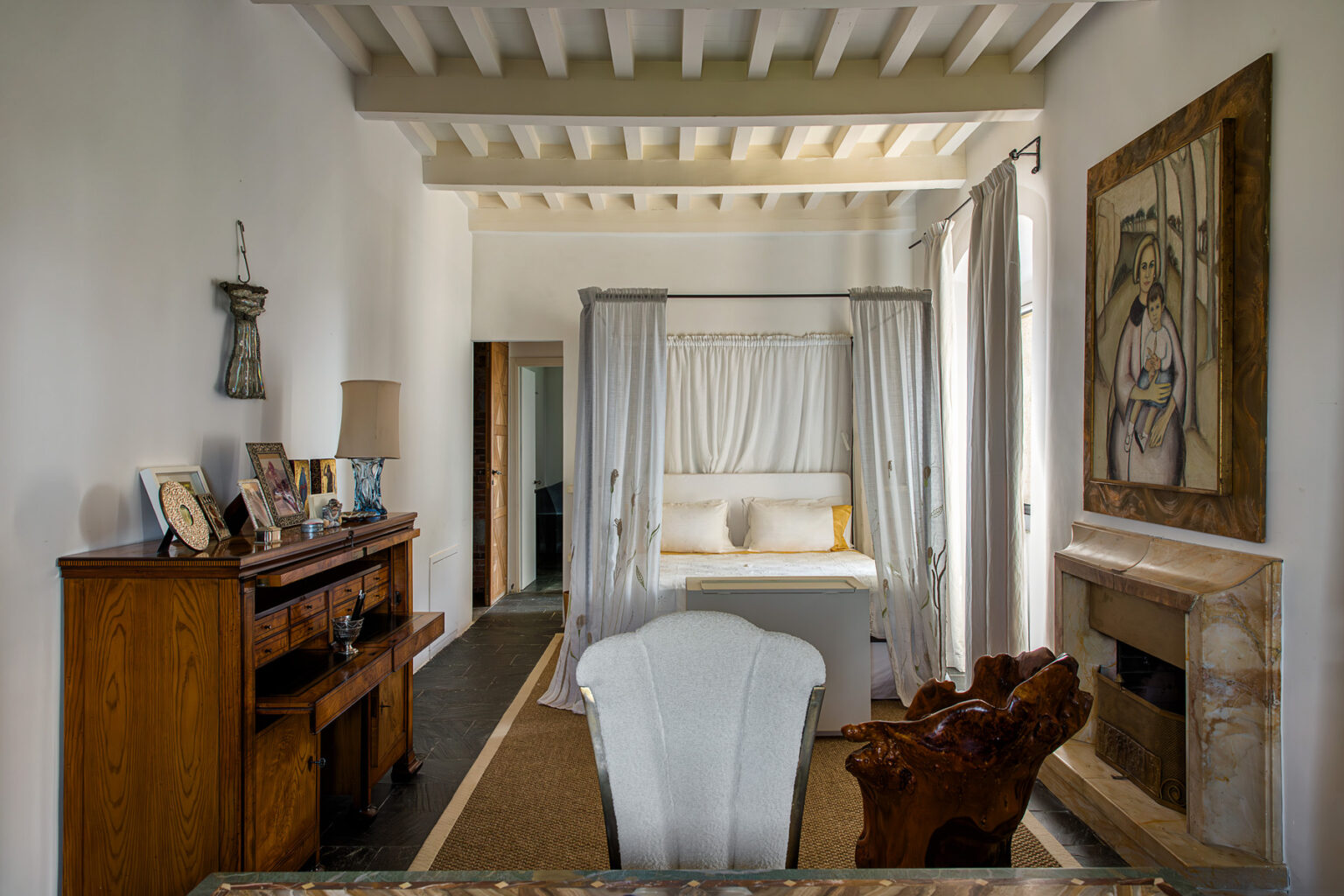 Interior view of a cozy bedroom featuring a four-poster bed with light drapes, a wooden dresser, and a decorative painting on the wall. The room has white walls and exposed wooden beams, creating a warm and inviting atmosphere. | Only on SopranoVillas