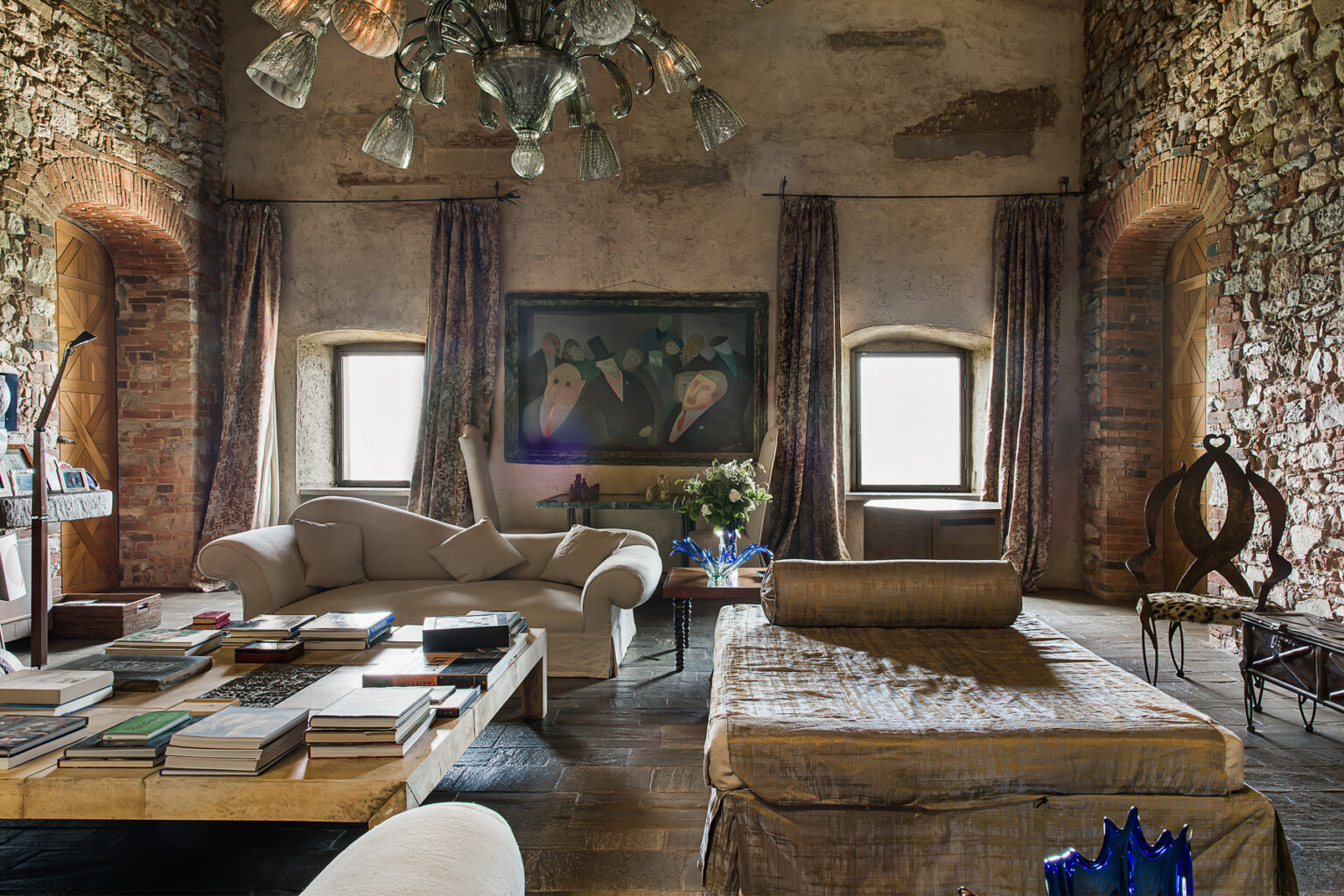 Spacious living room featuring rustic stone walls, elegant furniture, and large windows that provide natural light. The room includes a chandelier, a coffee table stacked with books, and decorative accents, creating a cozy yet sophisticated atmosphere. | Only on SopranoVillas