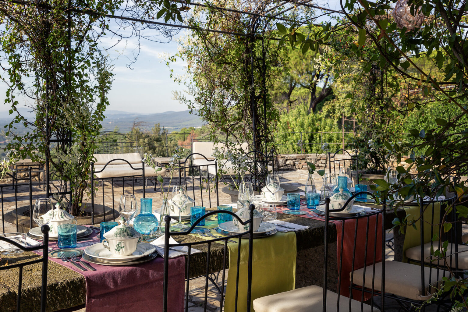 Elegant outdoor dining setup with a scenic view, featuring a long table adorned with colorful table linens, glassware, and plates, surrounded by lush greenery and vines. Ideal for al fresco dining experiences. | Only on SopranoVillas