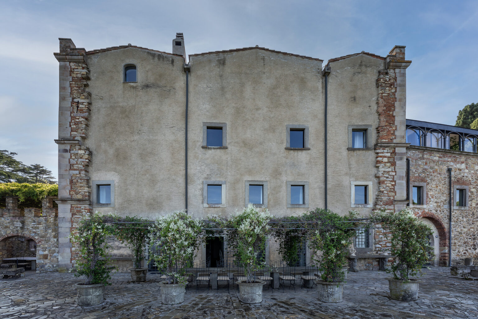 Historic building facade with stone and plaster elements, featuring multiple windows and a garden area with flowering plants in pots. The structure showcases a blend of architectural styles, set against a clear blue sky. | Only on SopranoVillas