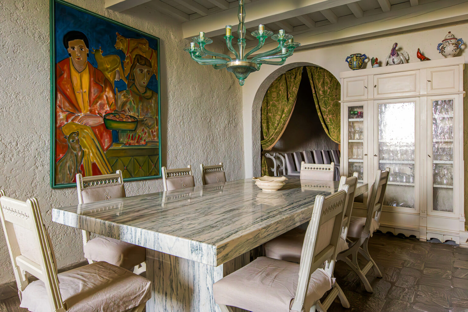 Elegant dining room featuring a large marble table surrounded by upholstered chairs, complemented by a colorful painting on the wall. A stylish chandelier hangs above, and a glass cabinet displays decorative items, contributing to the room's sophisticated ambiance. Green drapes frame a cozy nook in the background. | Only on SopranoVillas