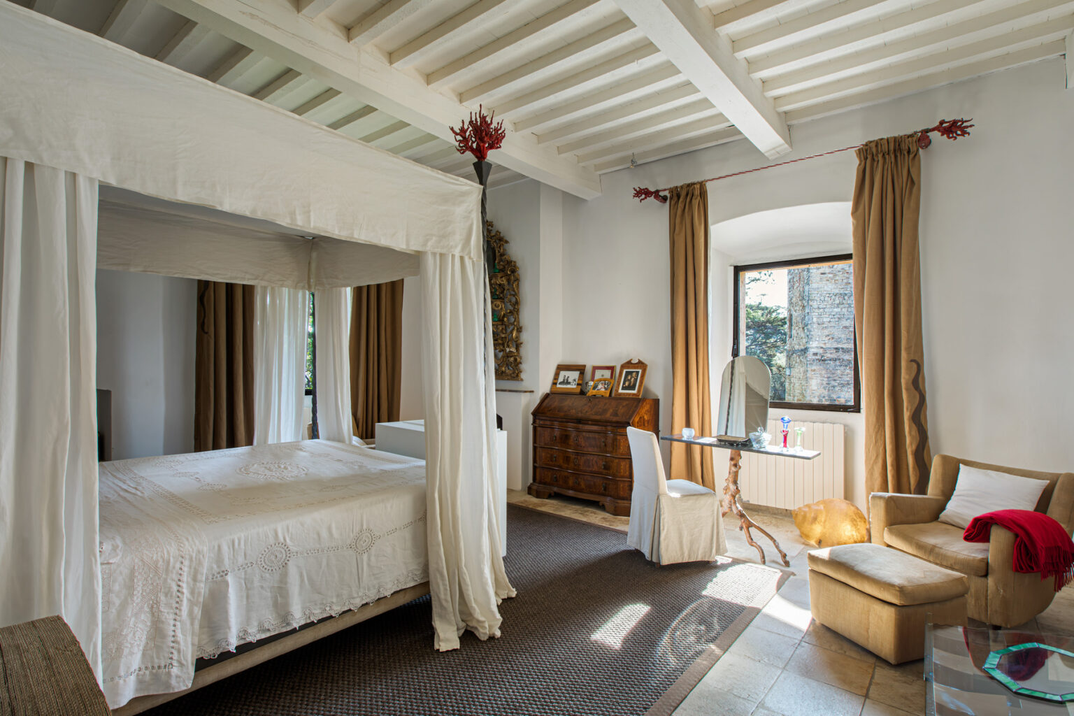 Spacious bedroom featuring a four-poster bed with white drapes, wooden furniture, and large windows with natural light. Decorated with warm curtains and a cozy seating area, the room exudes a tranquil and inviting atmosphere. | Only on SopranoVillas