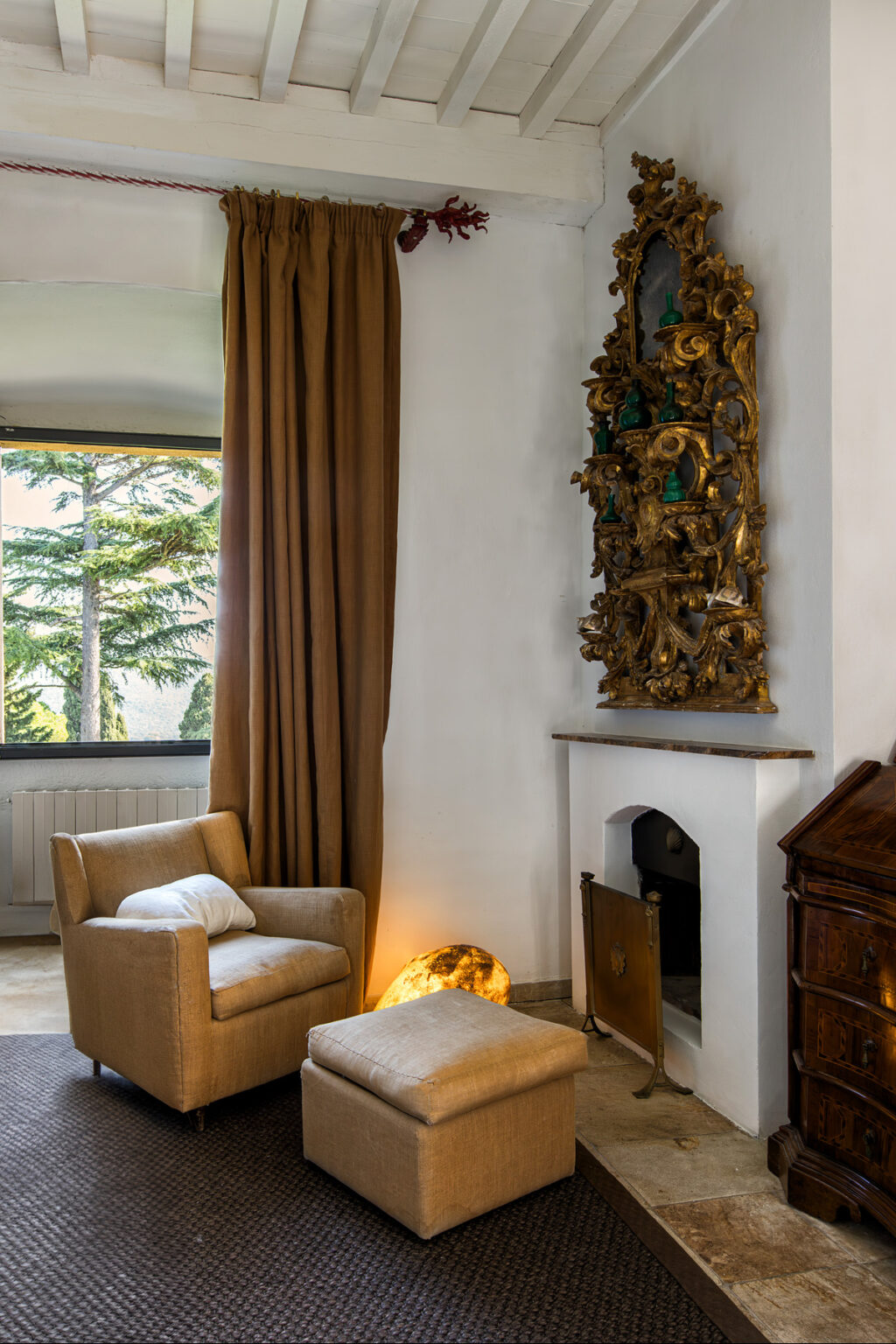Cozy living room featuring a beige armchair, elegant brown curtains, a decorative wall sculpture, and a classic fireplace, with large windows offering a view of trees outside. | Only on SopranoVillas