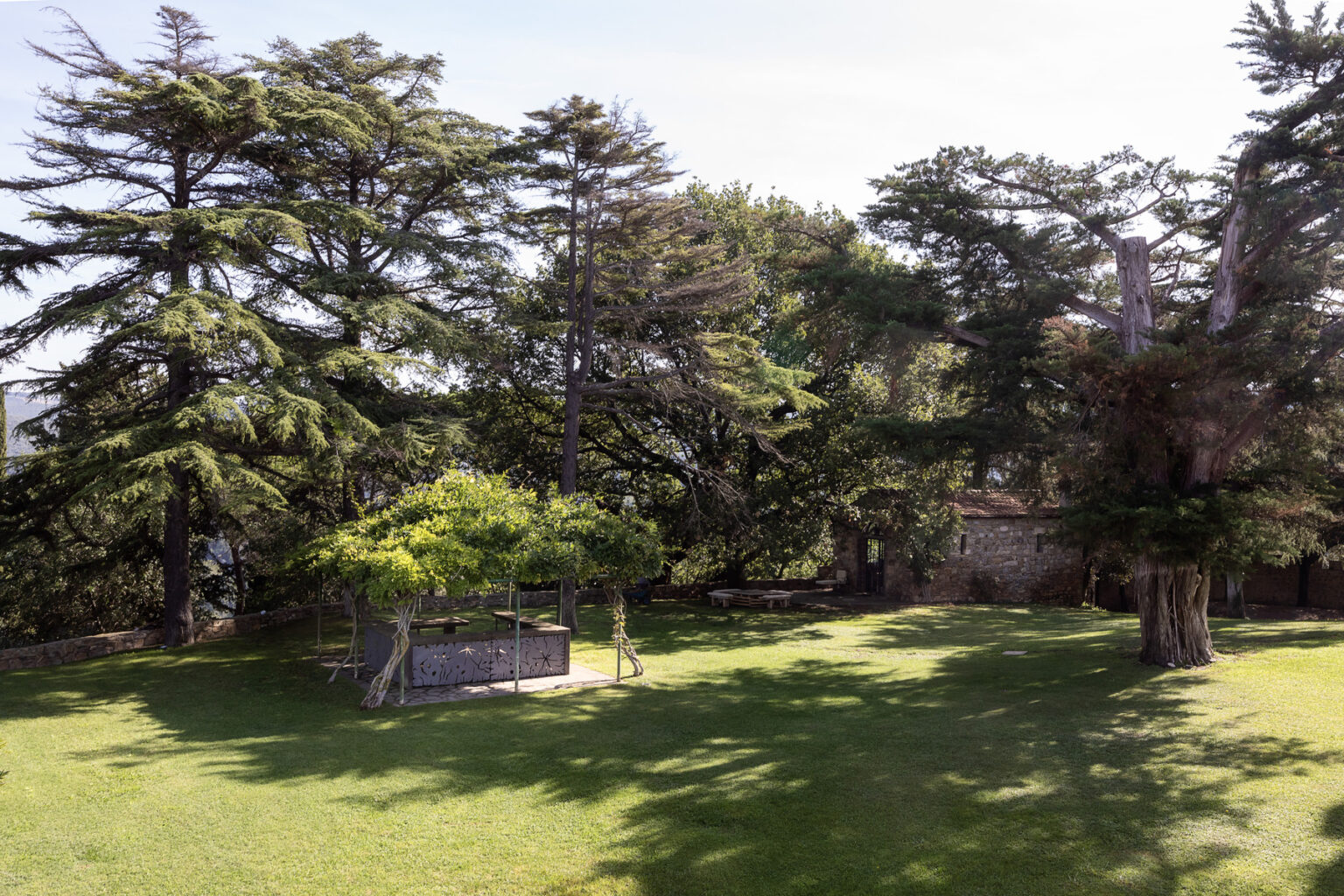 A scenic view of a lush green park featuring tall cedar trees, a shaded gazebo, and a well-maintained lawn under a clear blue sky. | Only on SopranoVillas