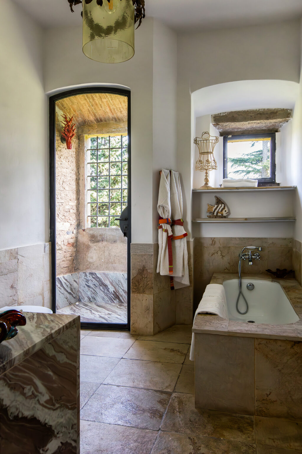 Modern bathroom design featuring a freestanding bathtub, marble surfaces, and a glass-enclosed shower. Natural light filters through a large window, highlighting the elegant decor, including a chandelier and decorative items on shelves. Soft towels and a robe add to the luxurious ambiance. | Only on SopranoVillas