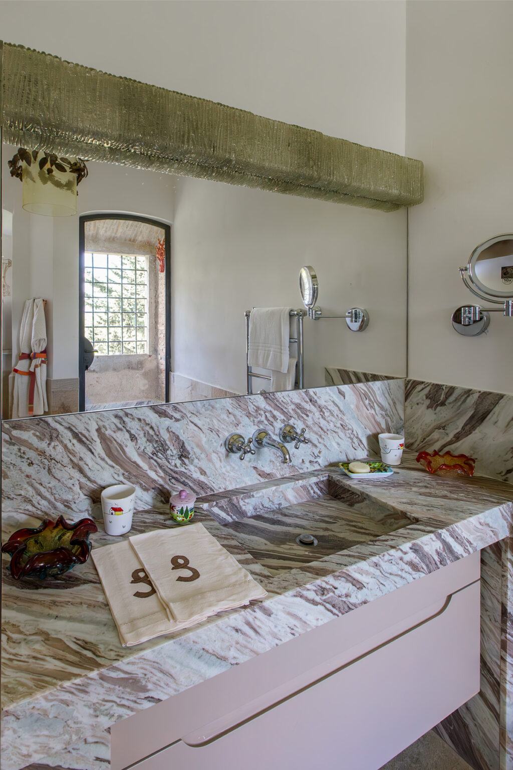 Modern bathroom featuring a marble countertop with dual faucets, neatly arranged towels, decorative bowls, and a large mirror reflecting natural light from a window. The design combines elegance with functionality, showcasing a blend of textures and colors. | Only on SopranoVillas