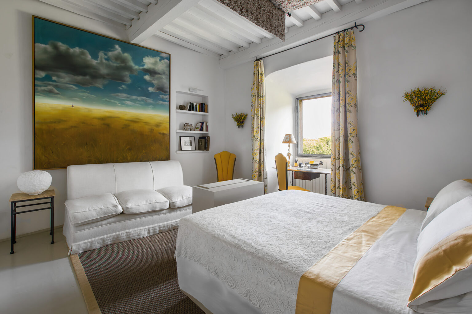 Bright and airy bedroom featuring a white bed with a decorative yellow trim, a cozy white sofa, and a wooden desk by a window. The room is adorned with floral curtains, a large landscape painting on the wall, and a stylish lamp, creating a serene and inviting atmosphere. | Only on SopranoVillas