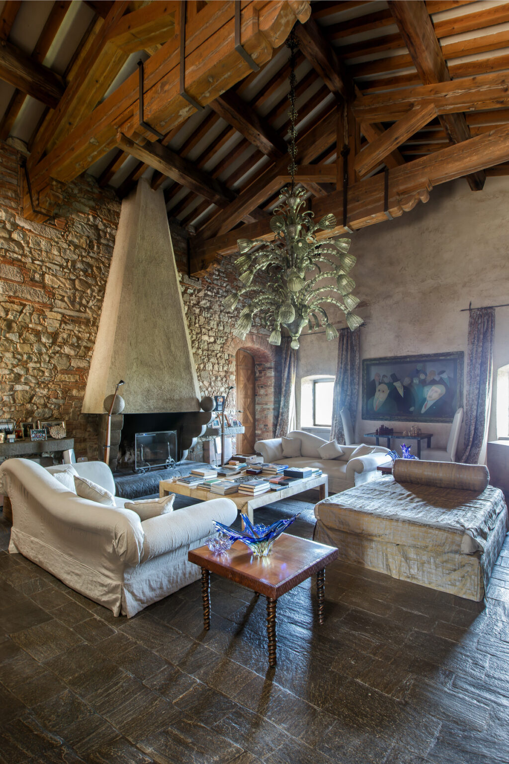 Cozy living room featuring a stone wall, a large fireplace with a modern chimney, plush white sofas, a wooden coffee table stacked with books, and elegant decor, including a striking chandelier and artwork on the walls. | Only on SopranoVillas