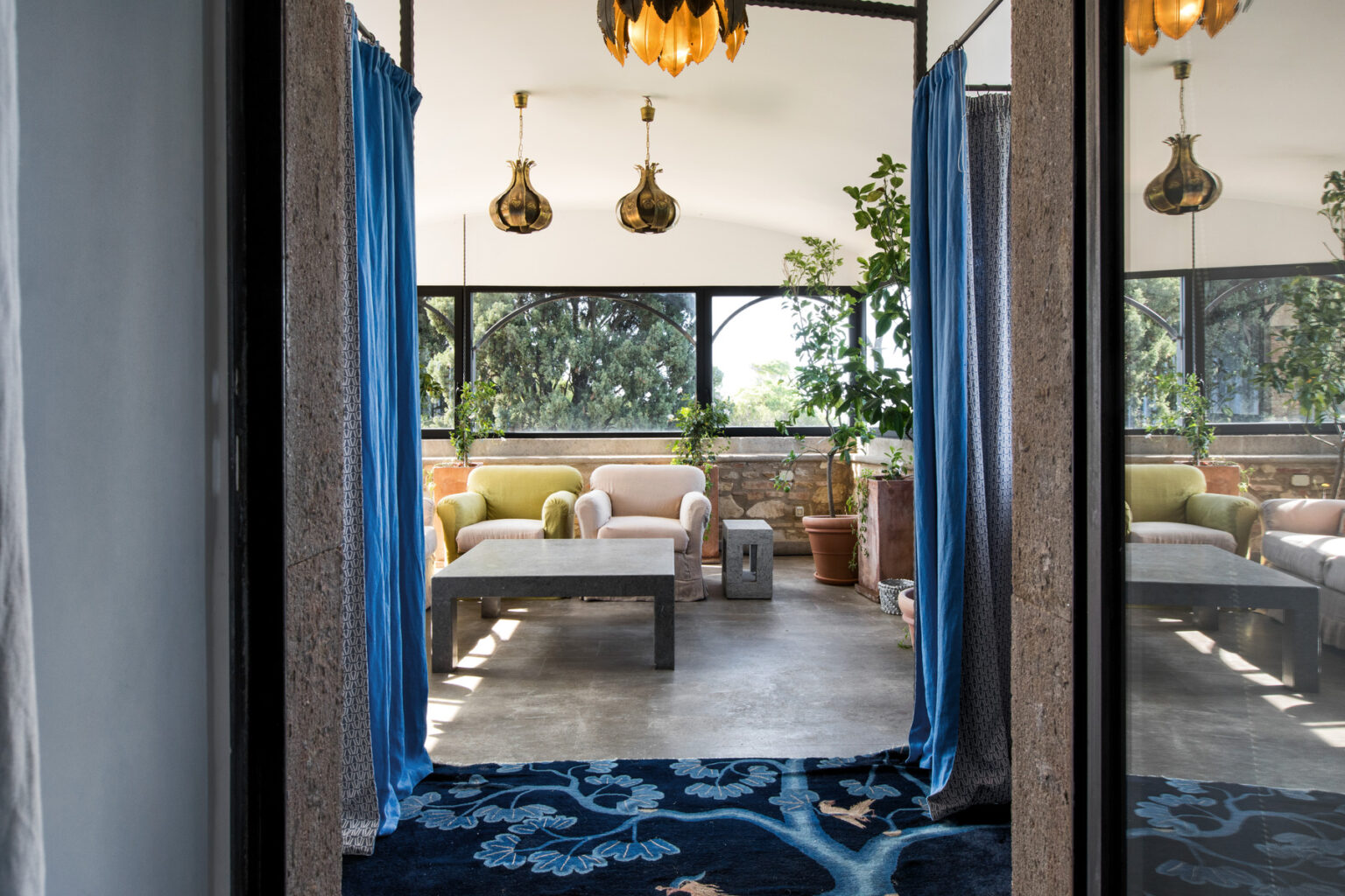 Bright and inviting interior view of a cozy living space featuring comfortable seating, potted plants, and large windows with natural light, framed by blue curtains and stylish pendant lighting. | Only on SopranoVillas