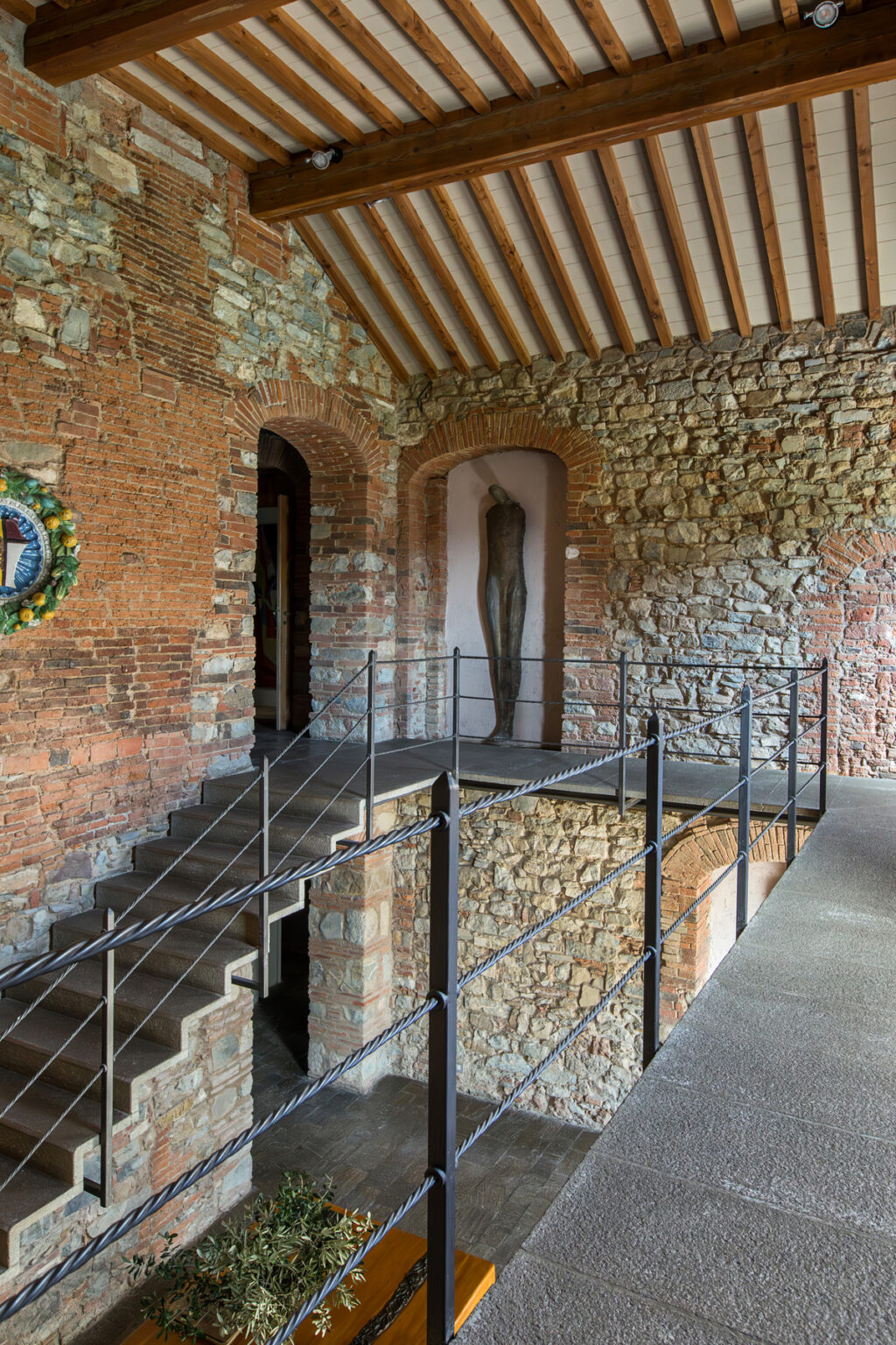 Interior view of a historic building featuring a staircase, iron railing, and a sculpture on display against a brick wall. The architectural details highlight a blend of stone and brickwork, creating an inviting atmosphere for visitors. | Only on SopranoVillas