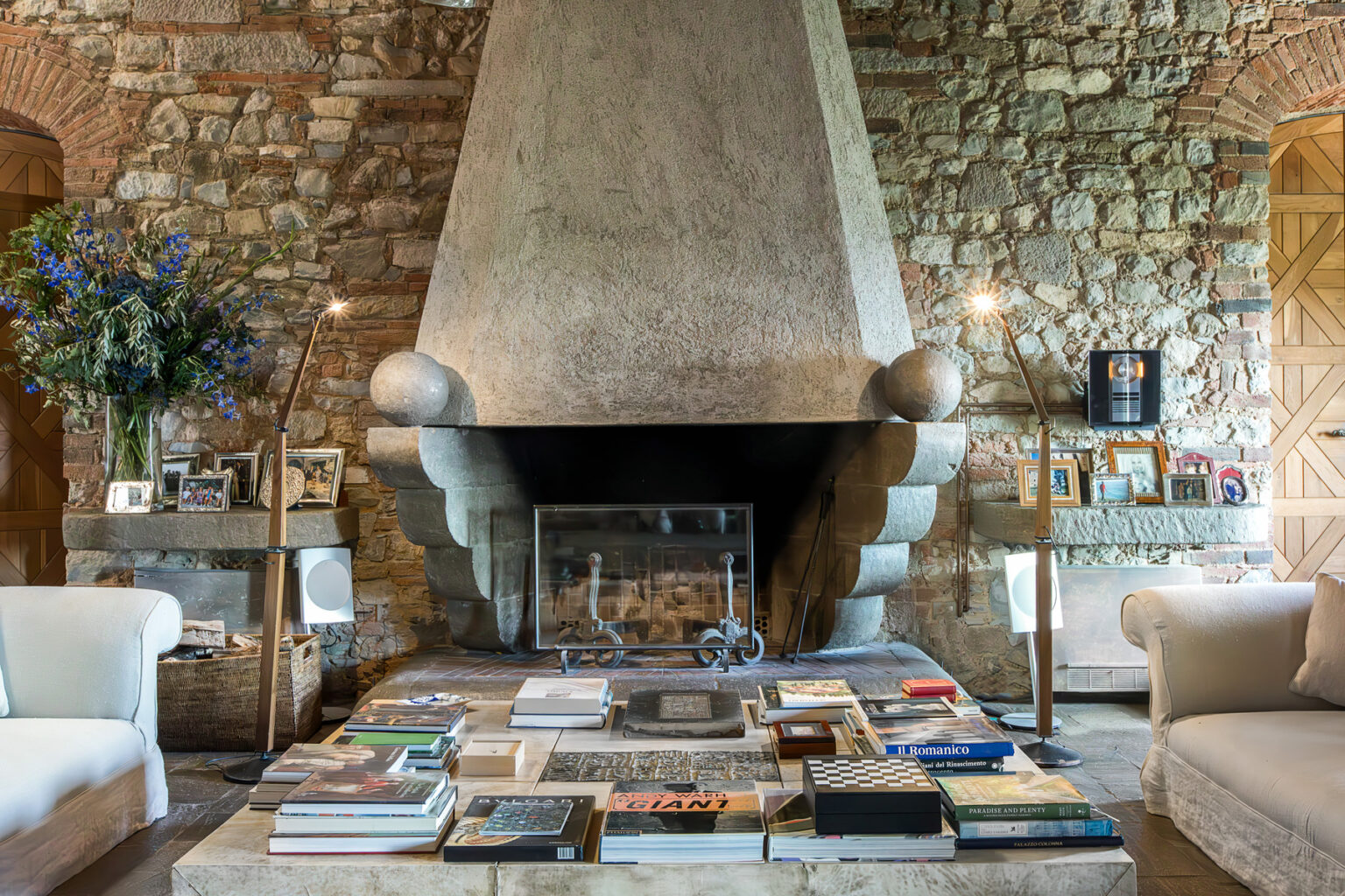 Cozy living room featuring a large stone fireplace with a unique design, surrounded by comfortable white armchairs, a coffee table stacked with books, and decorative elements like flowers and framed photographs. | Only on SopranoVillas