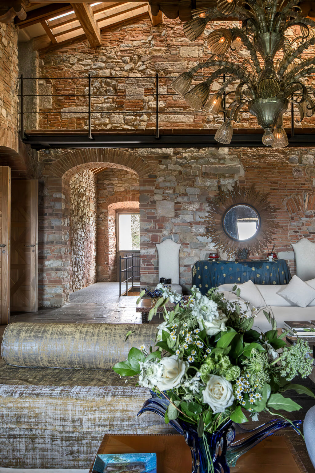 Interior of a rustic living space featuring exposed brick walls, a cozy seating area with a blue patterned throw, and a decorative mirror. Natural light filters through arched doorways, creating a warm and inviting atmosphere. | Only on SopranoVillas