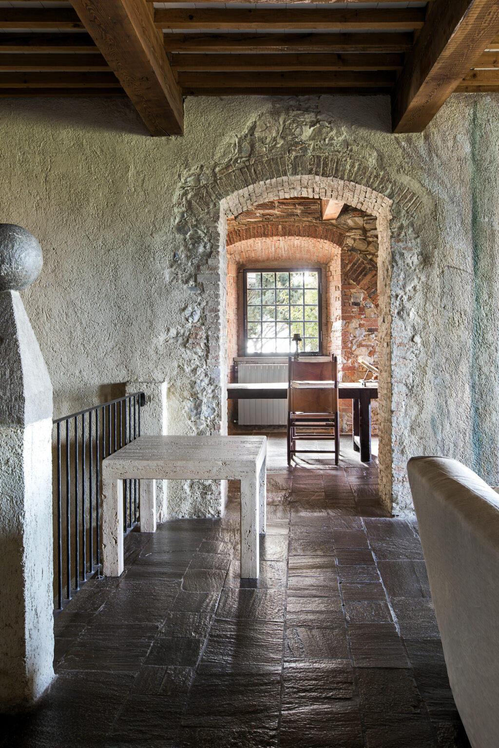 Interior view of a rustic room featuring a white minimalist table, stone flooring, and an arched doorway leading to a well-lit area with wooden furniture. The walls display a mix of textured plaster and exposed brick, complemented by wooden beams overhead. | Only on SopranoVillas