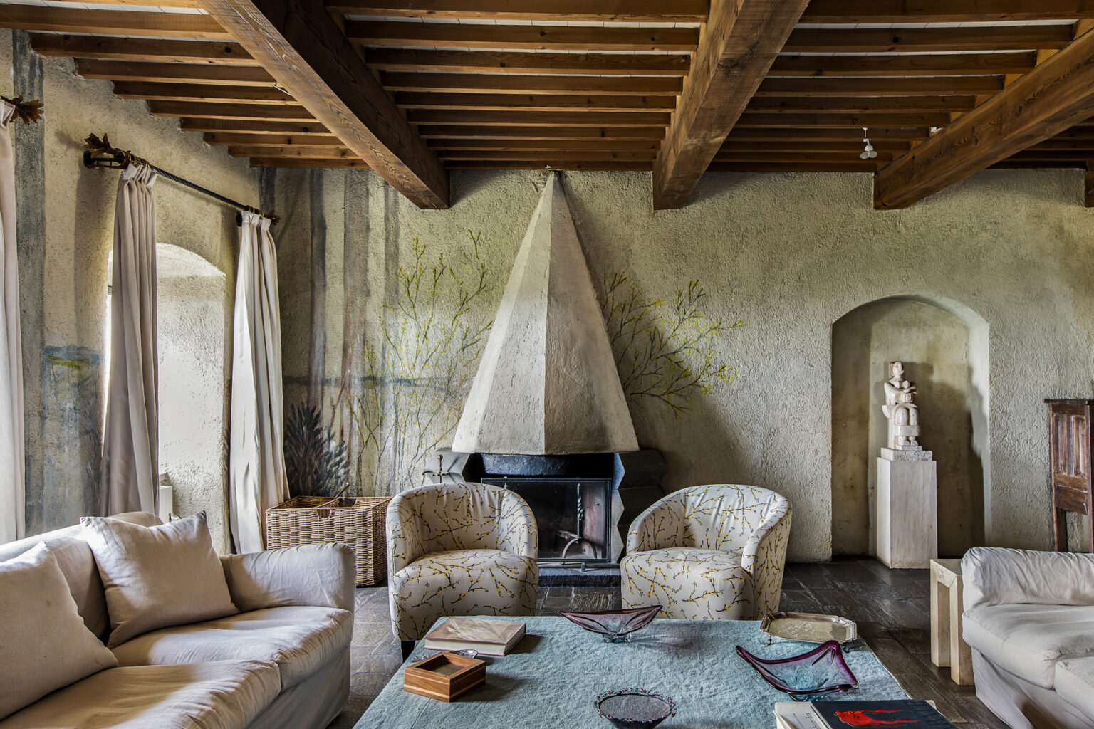 Cozy living room featuring rustic wooden beams, a unique pyramid-shaped fireplace, and comfortable seating. The space is adorned with soft textiles and artistic decor, creating a warm and inviting atmosphere. | Only on SopranoVillas