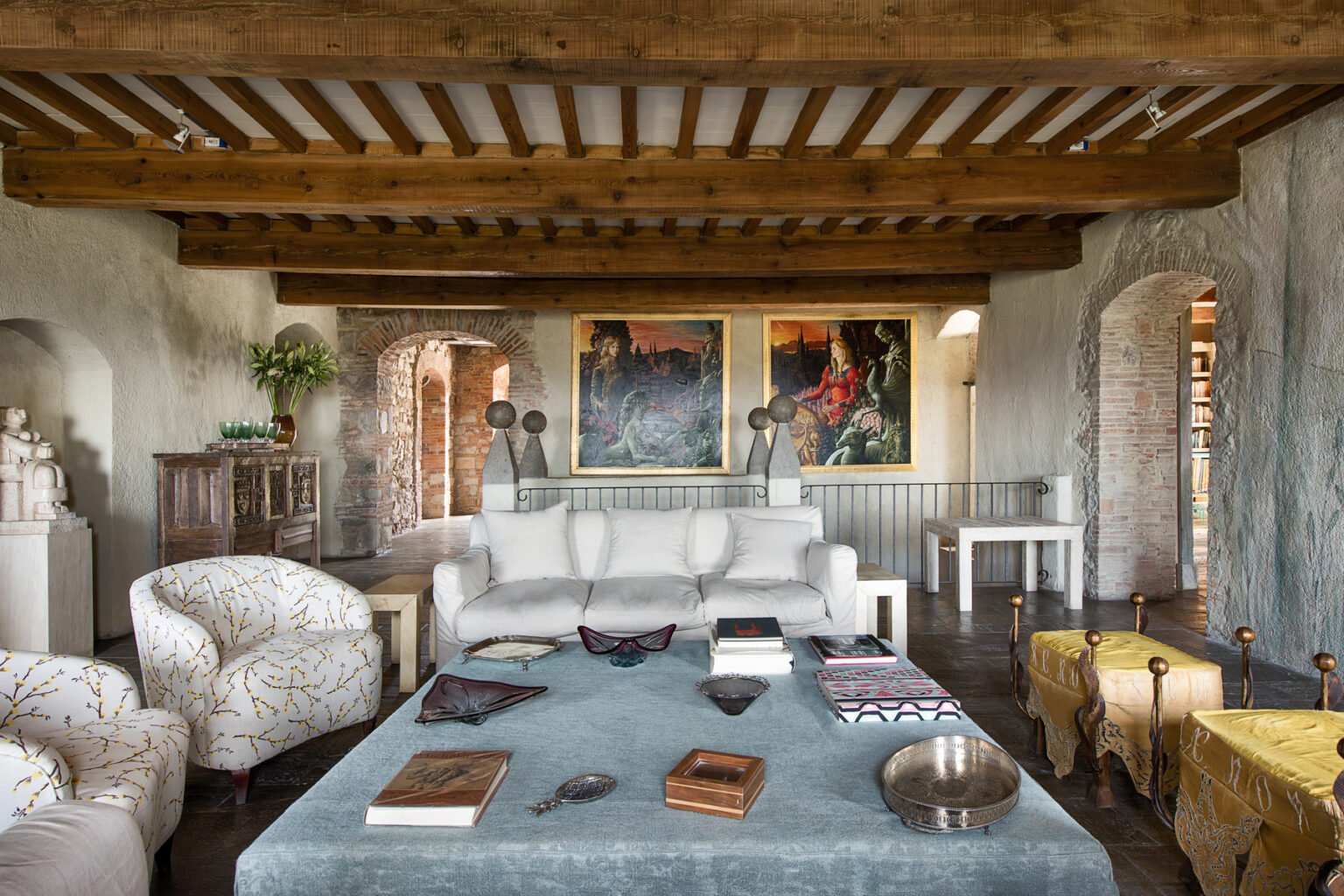 Cozy living room featuring a white sofa, two patterned armchairs, a wooden coffee table with books and decorative items, and vibrant artwork on the walls, all set under a wooden beam ceiling. | Only on SopranoVillas