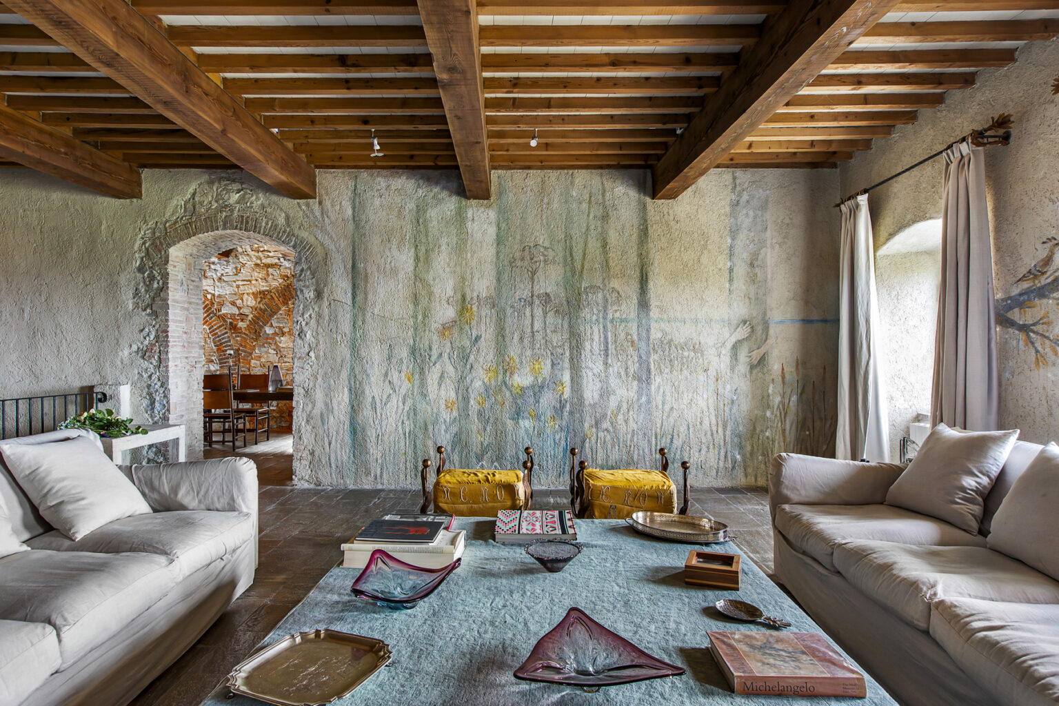 Cozy living room featuring a textured mural wall, wooden beam ceiling, and comfortable sofas with decorative cushions. The space is enhanced by natural light from large windows and a rustic coffee table adorned with books and decor items. | Only on SopranoVillas