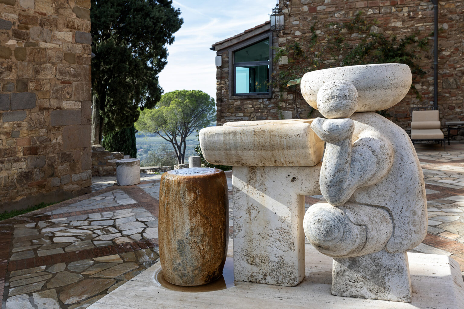 Sculpture of a seated figure made from light-colored stone, positioned next to a wooden barrel in an outdoor setting with stone walls and greenery in the background. | Only on SopranoVillas