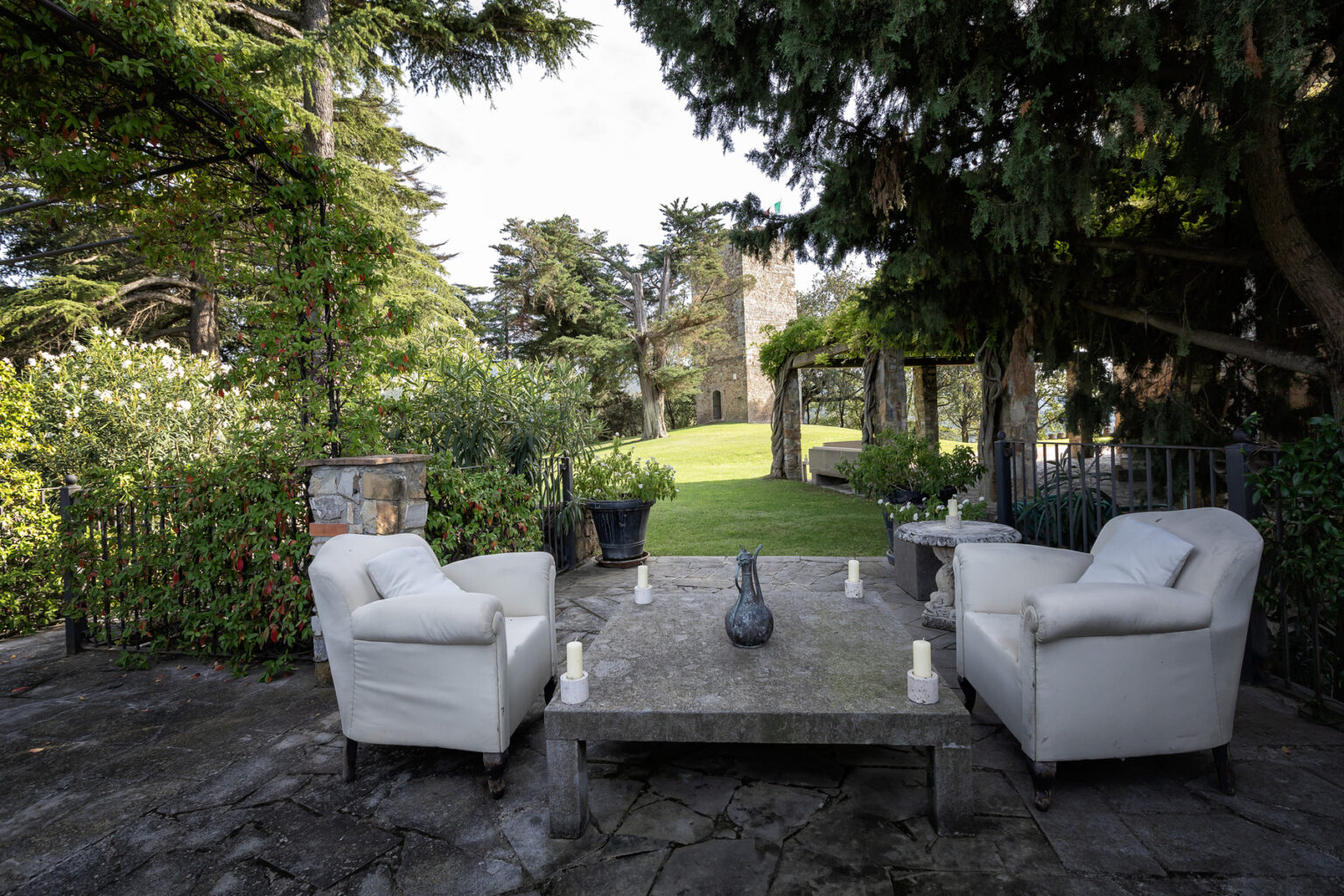 Cozy outdoor seating area featuring two white armchairs and a stone coffee table, surrounded by lush greenery and a well-maintained lawn in the background. Ideal for relaxation and gatherings in a tranquil setting. | Only on SopranoVillas