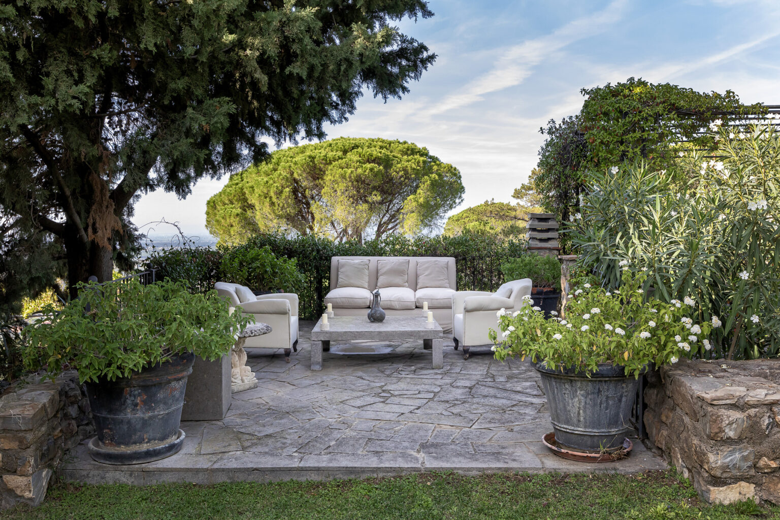 Outdoor seating area with a modern couch and armchairs surrounded by lush greenery and decorative planters, set against a scenic backdrop of trees and blue sky. | Only on SopranoVillas