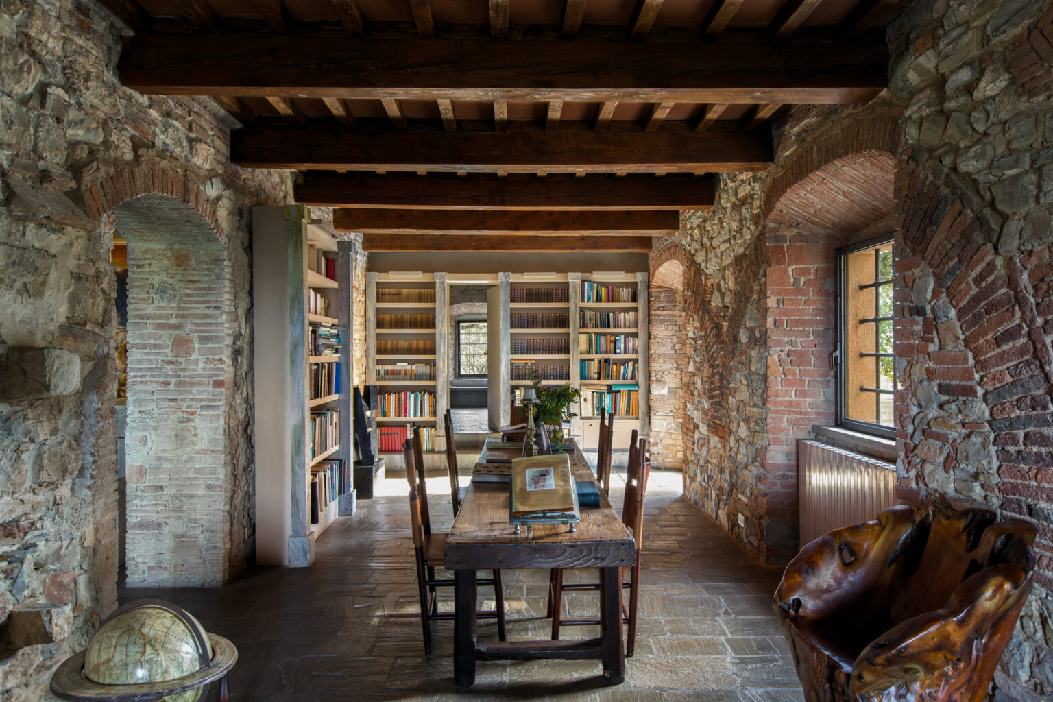 Cozy rustic interior of a library with exposed brick walls, wooden beams, and a large wooden dining table surrounded by chairs. Bookshelves filled with books line the walls, creating a warm and inviting reading space. A globe and decorative elements add character to the room. | Only on SopranoVillas