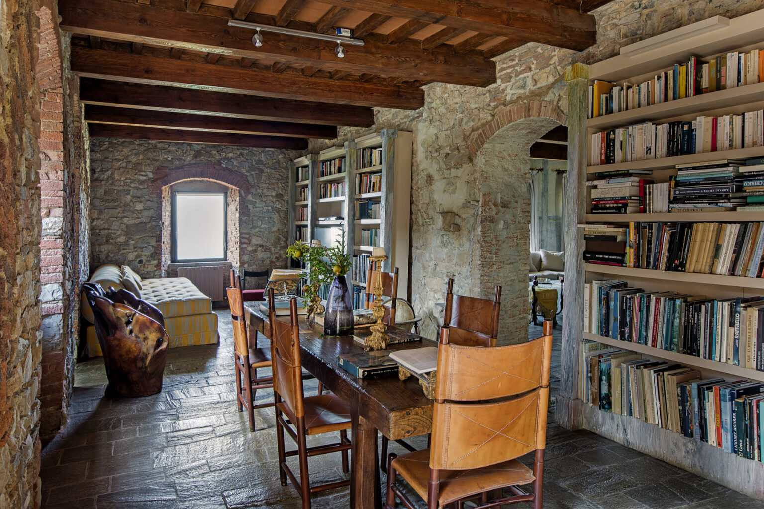 Cozy rustic interior of a library with wooden beams, a wooden dining table surrounded by chairs, a bookshelf filled with books, and a comfortable seating area with a view. | Only on SopranoVillas