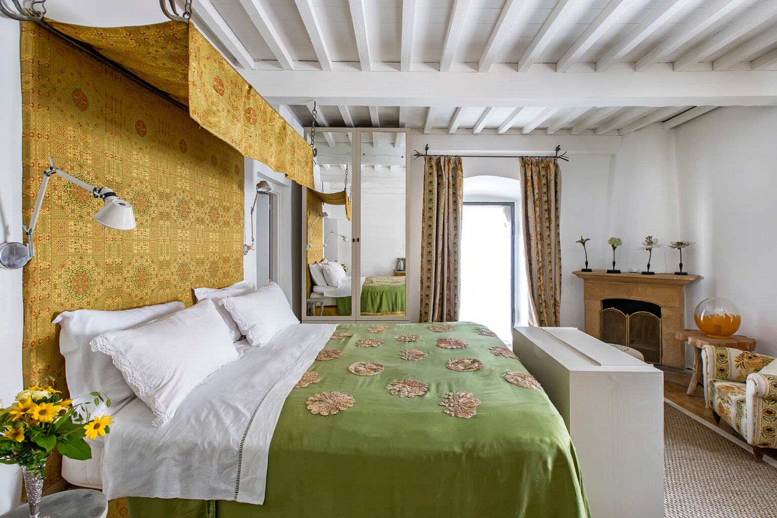 Cozy bedroom featuring a large bed with white linens and a green floral throw, complemented by a yellow patterned accent wall, elegant curtains, and a decorative fireplace, creating a warm and inviting atmosphere. | Only on SopranoVillas