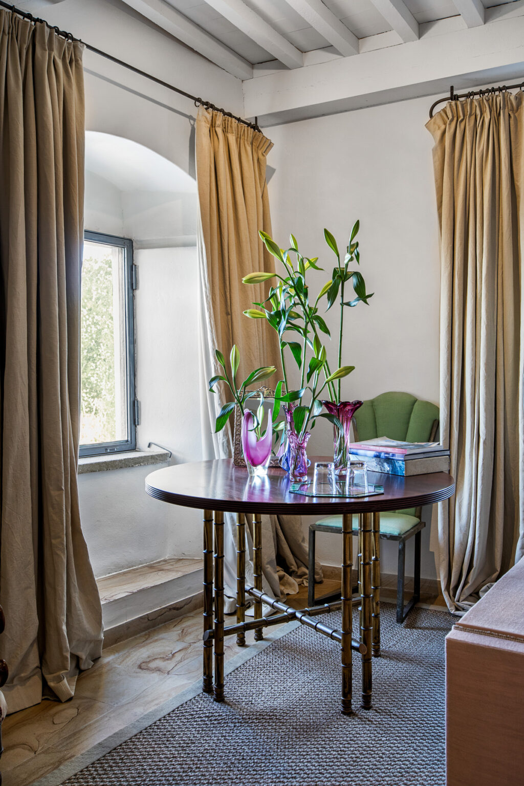 A cozy interior scene featuring a round wooden table adorned with colorful vases and fresh greenery, complemented by soft curtains and a comfortable green chair near a bright window. | Only on SopranoVillas