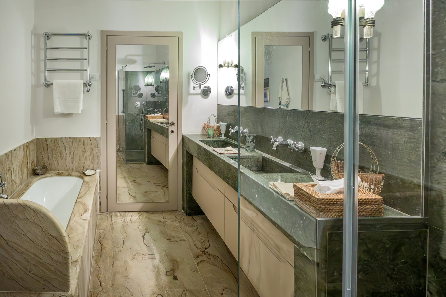 Modern bathroom interior featuring a sleek marble floor, double sink vanity with wooden cabinetry, a large mirror, and a glass shower enclosure. The space is illuminated by stylish wall sconces, creating a bright and inviting atmosphere. | Only on SopranoVillas
