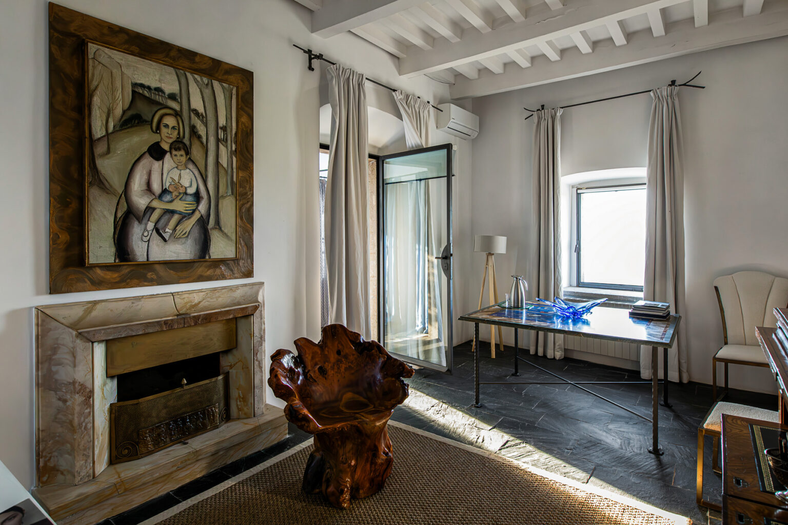 Interior view of a stylish living room featuring a modern art painting of a woman and child, a unique wooden chair, a glass-topped table with decorative items, and large windows with sheer curtains. The room has a marble fireplace and a cozy atmosphere, complemented by natural light and elegant furnishings. | Only on SopranoVillas