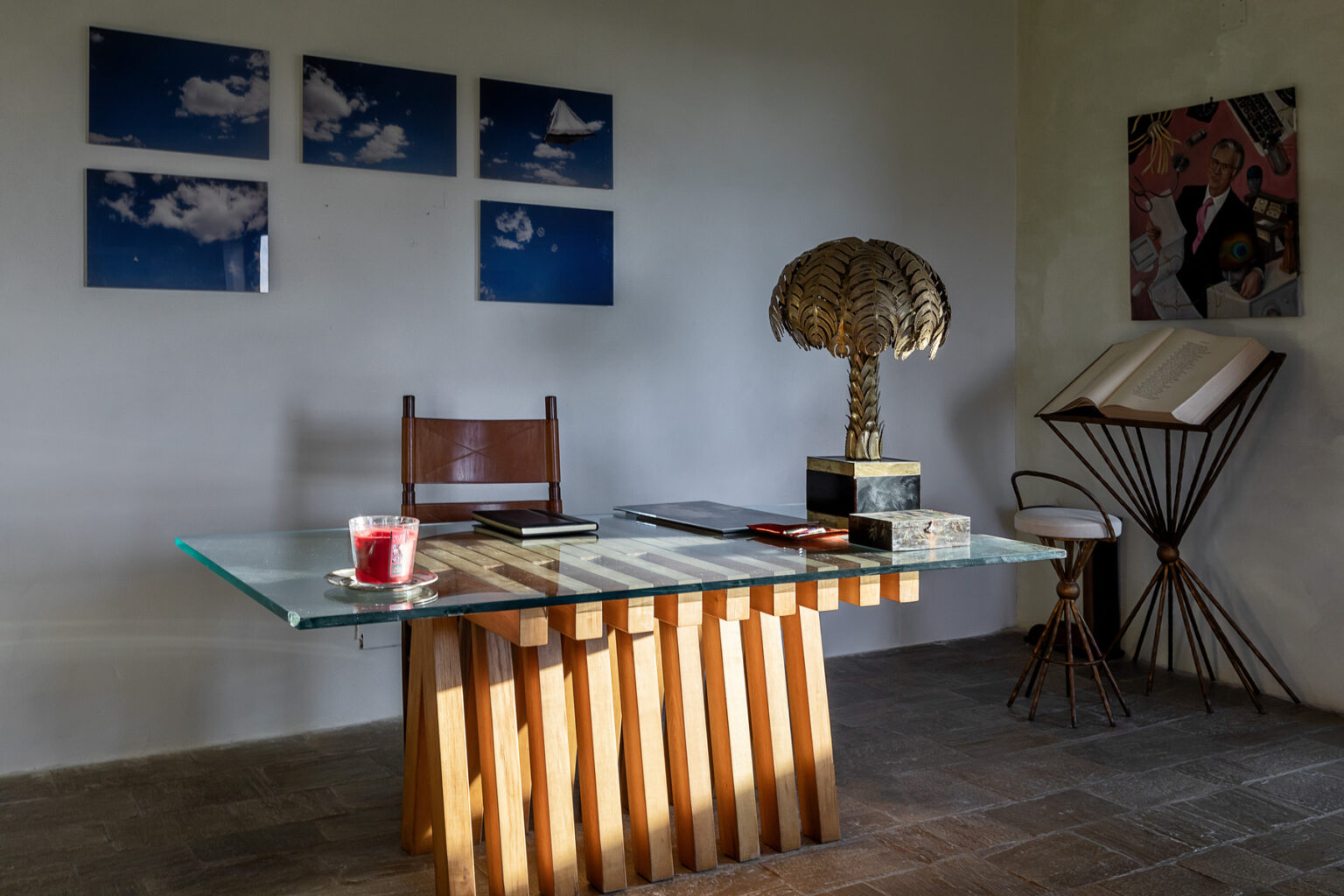 Modern office interior featuring a glass-top desk with a wooden base, a decorative lamp resembling a palm tree, and artistic wall decor. A leather chair and various notebooks are arranged on the desk, with a candle and a side table displaying an open book, creating a stylish and inviting workspace. | Only on SopranoVillas