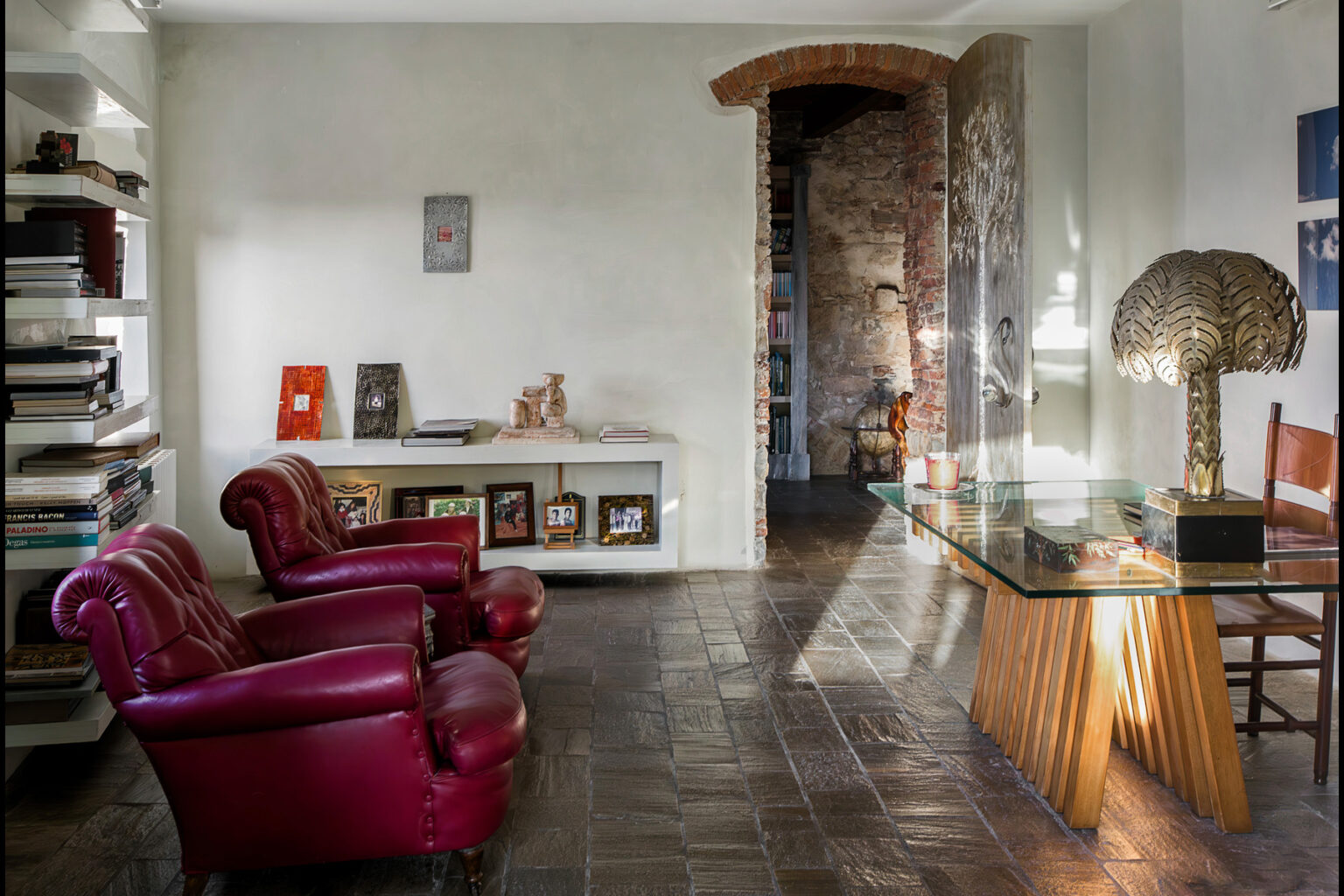 Modern living room interior featuring two red leather armchairs, a glass-top wooden table, and a decorative lamp. The room includes a bookshelf filled with books, framed photographs on display, and a rustic stone wall leading to another space. Natural light illuminates the area, enhancing the contemporary design. | Only on SopranoVillas
