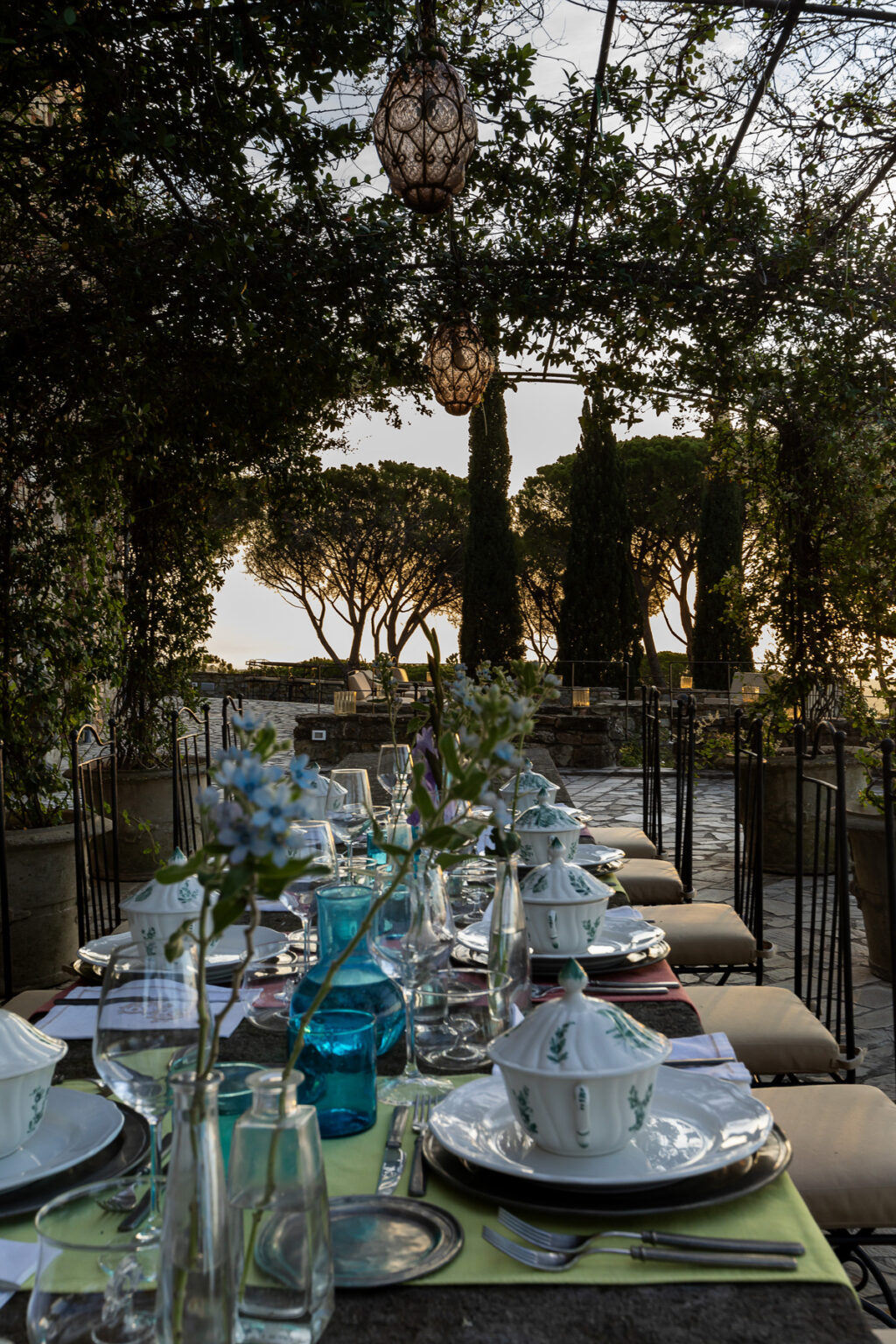 Outdoor dining setup featuring an elegantly arranged table with fine china, glassware, and floral centerpieces, surrounded by lush greenery and soft lighting, ideal for a romantic or special occasion. | Only on SopranoVillas